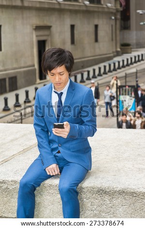 Man texting on street. Dressing in blue suit, necktie, a young Japanese Businessman sitting outside an office building in New York, looking down, checking messages on his mobile phone.