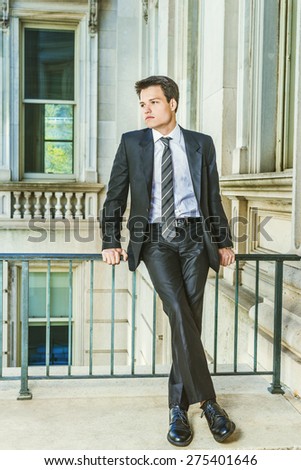 Dressing in black suit, necktie, white shirt, leather shoes, a young college student sitting by railing inside vintage style office building on campus, relaxing, thinking. Instagram filtered effect.