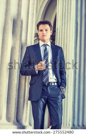 Young Businessman. Dressing formally in black suit, necktie, white shirt, wearing wristwatch, holding fist on chest, a young college student standing by column outside office, relaxing, thinking.