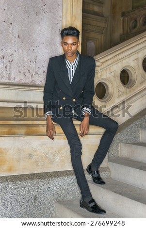School boy.  A young dark skin, 18 years old student, wearing black fashionable blazer, pants, leather shoes, standing by column on stairs inside vintage style office building, relaxing, thinking.