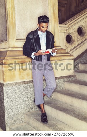 A young,18 years old student, wearing black jacket, striped pants, leather shoes, standing on stairs, leaning against column on campus, hands holding red book, reading. Instagram filtered effect.
