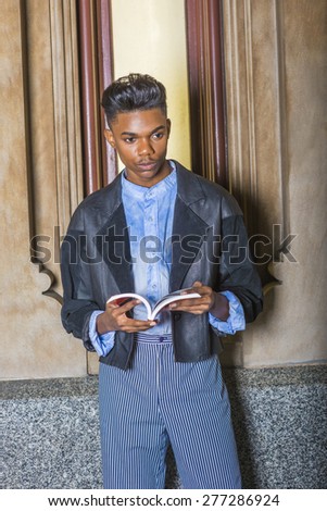 A young,18 years old student, wearing black fashionable jacket, blue dyed white shirt, striped pants, standing against small window on campus, hands opening book, reading, thinking, lost in thought.