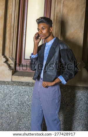Technology in Daily Life. A young, 18 years old student, wearing black fashionable jacket, blue dyed white shirt, striped pants, standing by window on campus, making phone call on his mobile phone.
