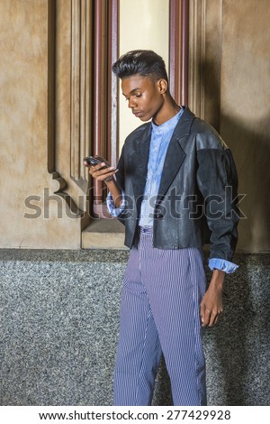 Technology in Daily Life. A young, 18 years old student, wearing black fashionable jacket, blue dyed white shirt, striped pants, standing by small window on campus, texting on his mobile phone.