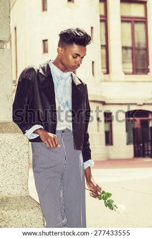 Lonely boy. A 18 years old student, wearing black fashionable jacket, striped pants, blue dyed white shirt, standing by gate on campus, holding white rose, lower hand, sad, thinking. Retro effect.