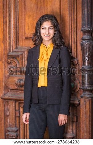 Portrait of Modern East Indian American Lady. Wearing black blazer, orange under shirt, a beautiful business woman with long curly hair standing by vintage style office door way, warmly greeting you.