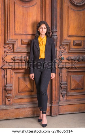 Graduate Student. Dressing in black blazer, orange under shirt, pants, heels, East Indian American business woman with long curly hair standing by vintage style office door way, warmly greeting you.