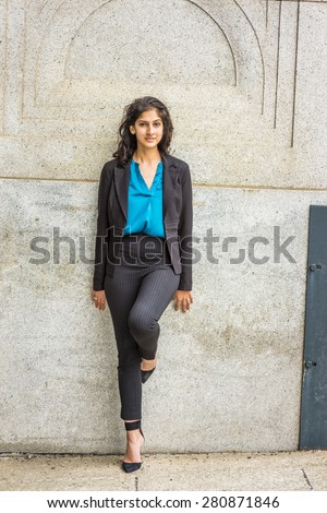 Modern East Indian American Student. Dressing in black blazer, blue under shirt, striped pants, heels,  a young girl with long curly hair standing against wall on campus, smiling, looking at you.