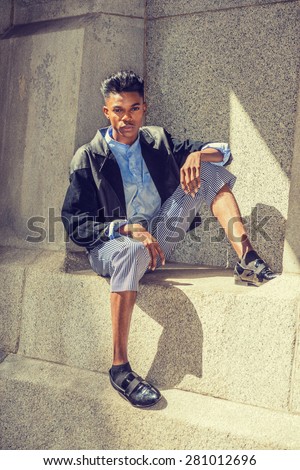 Urban teenage fashion. A 18 years old black student, wearing fashionable jacket, striped pants, blue dyed white shirt, leather shoes, sitting by rocky wall under sun shine, thinking. Instagram effect.