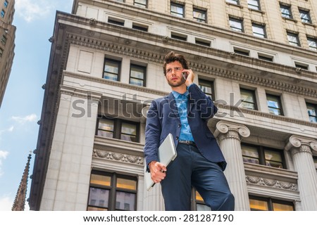 Power of Technology in our modern daily life. Dressing in blue suit, a young European Businessman with beard standing outside in New York, arm carrying laptop computer, talking on mobile phone.