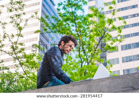 European graduate student studying in New York. Young guy with beard, sitting on the top of wall on business district in spring day, reading, working on laptop computer. Technology in daily life.