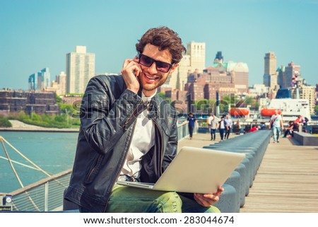 Businessman enjoying working outside. Wearing leather jacket, sunglasses, a guy with beard, sitting on bench at harbor, working on laptop computer, talking on phone in the same time. Instagram effect.