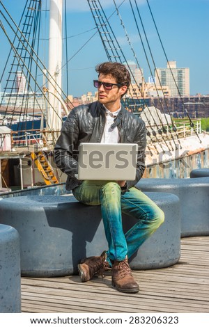 Traveling, working in New York. Wearing black leather jacket, jeans, brown boot shoes, sunglasses, a young guy with beard, sitting on bench at harbor, working on laptop computer. A boat on background