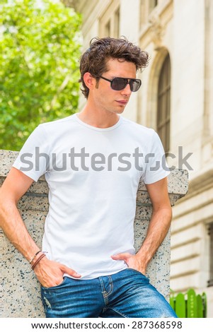 Man Summer Casual Street Fashion. Wearing White T shit, jeans, sunglasses, hands in pockets, a young European college student standing at corner on campus in New York, looking down, sad, thinking.
