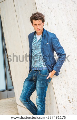 Man Urban Casual Fashion. Wearing blue blazer, patterned under shirt, jeans, hands in back pockets, a young European college student standing by column, looking down, sad, thinking, lost in thought.