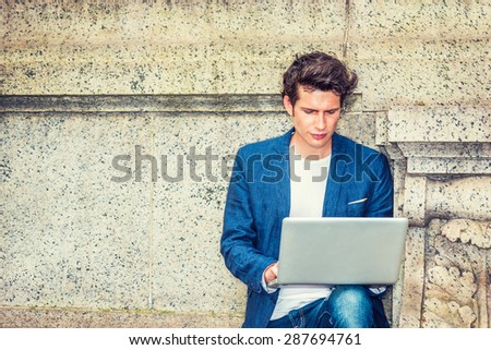 European college student studying outside on campus.  A young businessman sitting against wall, looking down, reading, thinking, working on laptop computer. Technology in our daily life. Copy Space.