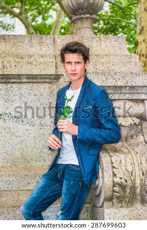 European college student seeking for love in New York. Wearing blue blazer, while under shirt, jeans, a young guy standing at corner against wall on campus, holding white rose, waiting for you.