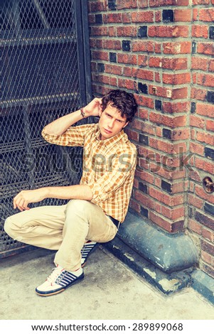 Concept of lonely man thinking about love. Wearing yellow patterned shirt, pants, sneakers, a young European guy squatting at corner on street in New York, sad, thinking after busy working day.