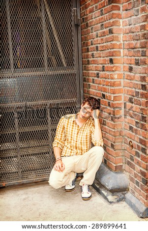 Concept of lonely man thinking about love. Wearing yellow patterned shirt, pants, sneakers, a young guy squatting at corner on street in New York, looking down, sad, thinking after busy working day.
