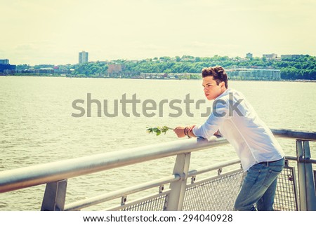 Man Missing You. Wearing white shirt, jeans, holding white rose, a guy standing by Hudson River in New York, opposite New Jersey, thinking, lost in thought. Concept of looking for love, friendship.