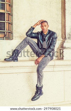 Man Urban Casual Fashion. Wearing black leather jacket, striped undershirt, jeans, leather boots, a young African American guy sitting against wall on street in New York, relaxing, waiting for you.