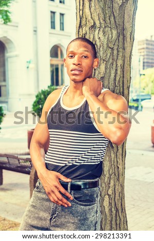 Summer Heat. Man casual fashion. Wearing black, white striped tank top, jeans, a young, sexy African American guy leaning against tree trunk on street, relaxing, thinking, lost in thought.