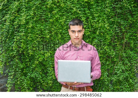 College student studying in peaceful, green environment. Wearing red patterned, long sleeve shirt,  a young guy standing against wall with green leaves on campus, reading, working on laptop computer.