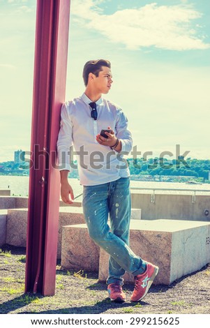Where are you? Wearing white shirt, jeans, sneakers, sunglasses hanging on collar, young guy standing against pole by Hudson River in New York, holding mobile phone, waiting for you. Instagram effect.