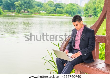 Working hard in peaceful environment. Wearing black suit, red, white patterned undershirt, a young college student sitting by lake with green trees on campus, reading, working on laptop computer.