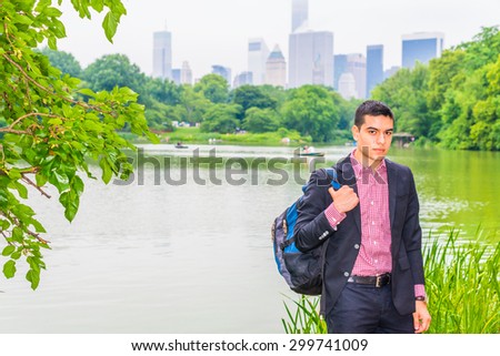 Wearing black blazer, red, white patterned under shirt, carrying shoulder bag, young college student standing by green lake in big city, confidently looking at you. Concept of Environment Protection.
