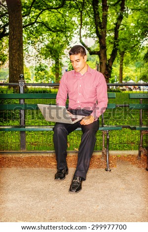 Man working outside. Wearing red, white patterned shirt, black pants, leather shoes, a young college student sitting on chair under trees, reading, working on laptop computer. Technology in daily life