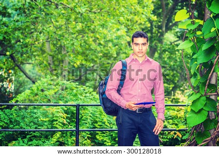 Back to School. Wearing patterned shirt, carrying shoulder bag, hand holding tablet computer, a college student standing by woods on campus, confidently looking forward. Fog, drizzle on background.