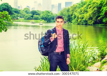 Wearing black blazer, red, white patterned under shirt, carrying shoulder bag, young college student standing by green lake in big city, confidently looking at you. Concept of Environment Protection.