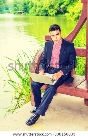 Working hard in peaceful environment. Wearing black suit, red, white patterned undershirt, leather shoes, a young college student sitting by lake on campus, working on laptop computer, thinking.