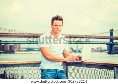 American Businessman working in New York. Wearing white T shirt, a young sexy college student  standing at harbor, reading, studying on laptop computer. Manhattan, Brooklyn bridges on background.