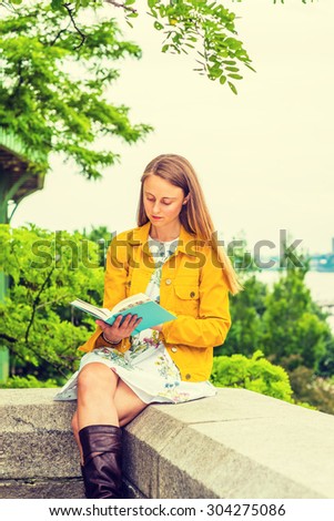 American college student studying in New York. A pretty girl wearing flower patterned underdress, yellow corduroy jacket, holding green book, sitting by trees on campus, looking down, reading.