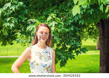 Summer Green. Wearing flower patterned, sleeveless, white dress, an American college student standing on green lawn by trees on campus, smiling, looking at you. Concept of Environment Protection.