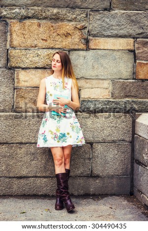 Portrait of College Student. A girl wearing flower patterned, sleeveless, white dress, brown leather high riding boots, holding green book, crossing legs, standing by rocky wall. Instagram effect