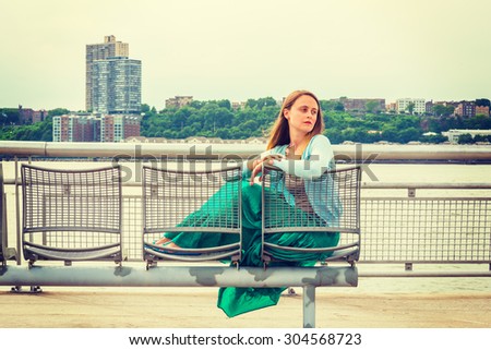 Lonely girl missing you. Wearing light blue sweater, green skirt, an American woman sitting on metal chair by Hudson River in New York, opposite New Jersey, looking around, thinking. Instagram effect
