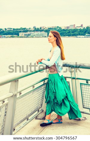 I waiting for you. American college student wearing light blue cardigan, green skirt, sandals, standing by metal fence by Hudson River in New York, opposite New Jersey, holding book, looking forward.