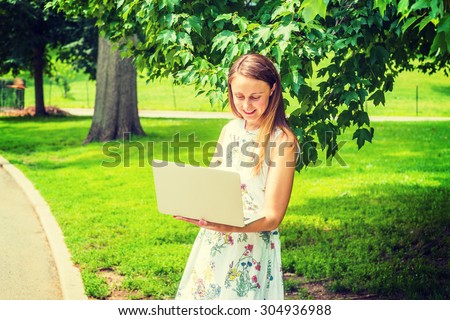 Technology in daily life. Wearing flower patterned white dress, a college student standing on green lawn by trees on campus, reading, working on laptop computer. Concept of Environment Protection.