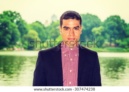Wearing black blazer, red, white patterned under shirt, a young college student standing by green lake on campus, confidently looking at you. Concept of Environment Protection. Instagram effect.