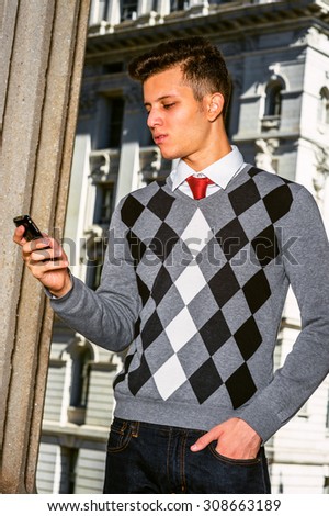 Technology in our daily life. Wearing black, white, gray patterned sweater, red necktie, jeans, a young college student checking messages on his mobile phone on street. Instagram filtered effect.