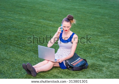 Busy Woman working hard. A young college student wearing polkadot dress, carrying leather bag, sitting on green lawn, working on laptop computer, unhappy, puzzled, making phone call for help.