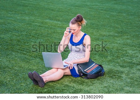 Busy Woman working hard. A young college student wearing polkadot dress, carrying leather bag, sitting on green lawn on campus, reading, working on laptop computer, talking on phone in the same time.