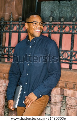 African American college student studying in New York. Dressing in blue shirt, brown pants, wearing glasses, holding laptop computer, young man standing against window on street, smiling, looking away