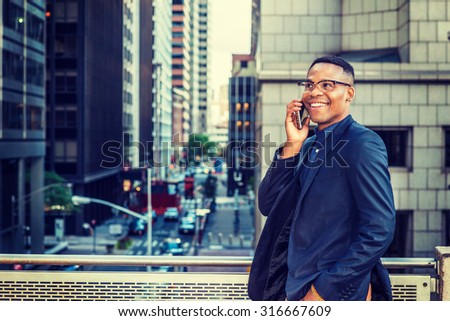 Happy African American Businessman working in New York. Wearing blue jacket, glasses, a young black man standing by railing on balcony, facing street with high buildings, smiling, talking on phone.