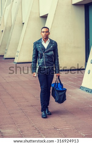 Man Urban Casual Fashion. Wearing black leather jacket, jeans, leather shoes, glasses, holding laptop computer, carrying duffel bag, an African American college student walking on street in New York.