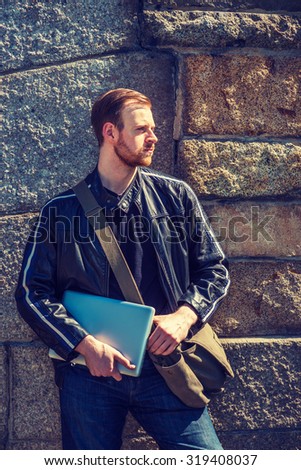 Wearing black leather jacket, jeans, carrying shoulder bag, holding laptop computer, an American man with beard, mustache standing by rocky wall, ready to work. Instagram effect.