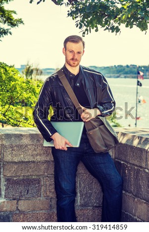 Wearing black leather jacket, jeans, carrying shoulder bag, holding laptop computer, young American man with beard, mustache standing by river, traveling, working in New York. Instagram effect.